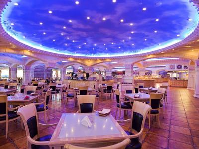 Amplio comedor de hotel con sillas modernas, mesas blancas, iluminación lila y techo decorativo.