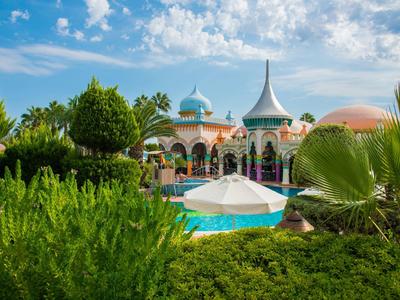 Una piscina frente a un edificio colorido con techos puntiagudos rodeado de árboles bajo un cielo azul.