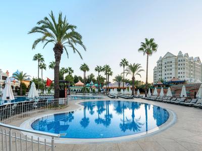 Rundes Schwimmbecken mit Sonnenschirmen und Palmen neben einem Hotelgebäude bei klarem Himmel.