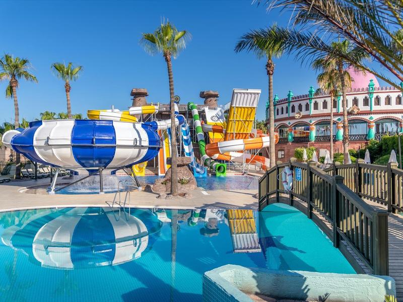 Tobogán acuático colorido y piscina con palmeras y cielo azul en un resort vacacional.