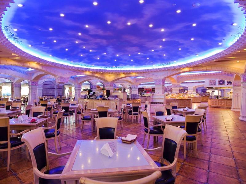 Amplio comedor de hotel con sillas modernas, mesas blancas, iluminación lila y techo decorativo.