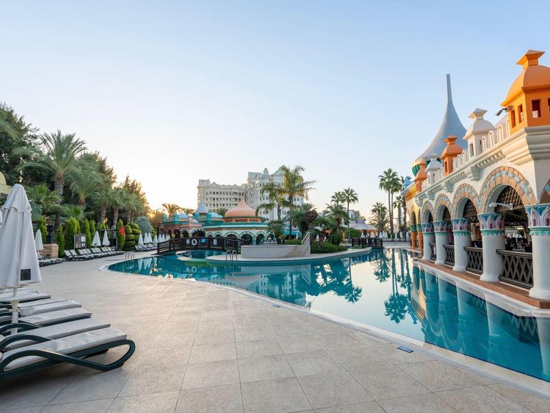 Piscina del hotel con tumbonas y palmeras, edificio con arcos y cielo despejado al atardecer.