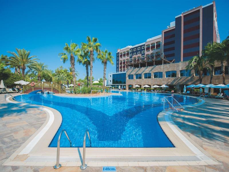 Gran piscina de hotel con palmeras, tumbonas y sombrillas bajo un cielo azul claro.