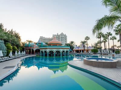 Basen hotelowy otoczony palmami, leżakami i wielopiętrowym budynkiem w tle.