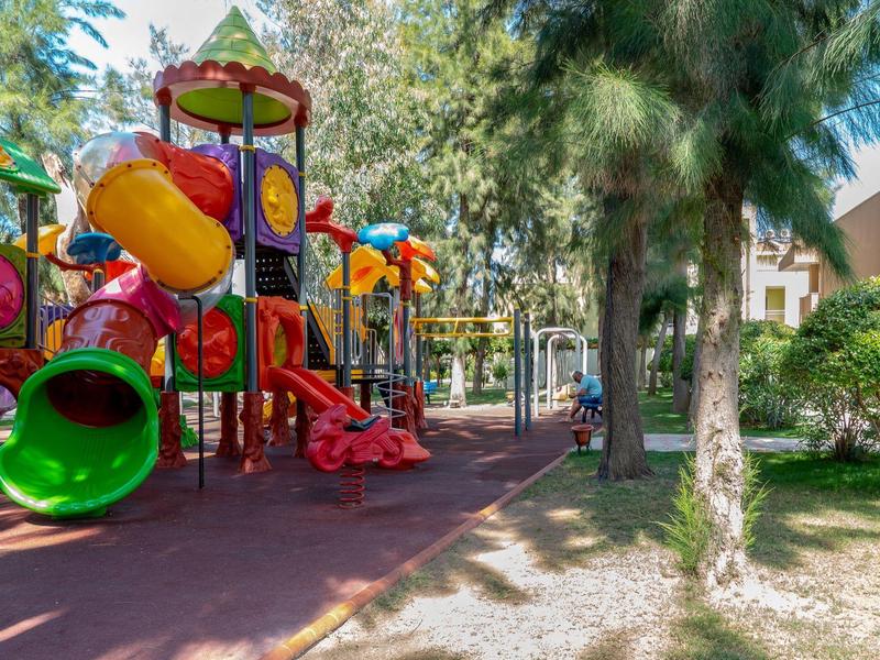 Kolorowy plac zabaw z zjeżdżalniami i sprzętem do wspinaczki otoczony drzewami obok hotelu.