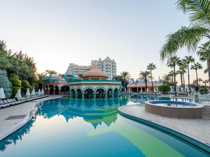 Basen hotelowy otoczony palmami, leżakami i wielopiętrowym budynkiem w tle.