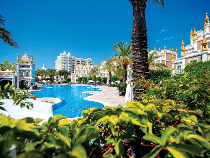 Widok na basen hotelowy z palmami i budynkami w stylu śródziemnomorskim w tle.