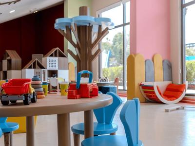 Helles Kinderspielzimmer mit bunten Möbeln, einem großen Fenster und Spielzeugen auf einem Tisch.