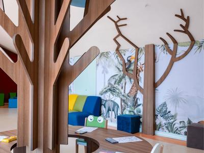 Kinderspielbereich mit stilisiertem Baum aus Holz, Stühlen und Malutensilien in einem hellen Raum.