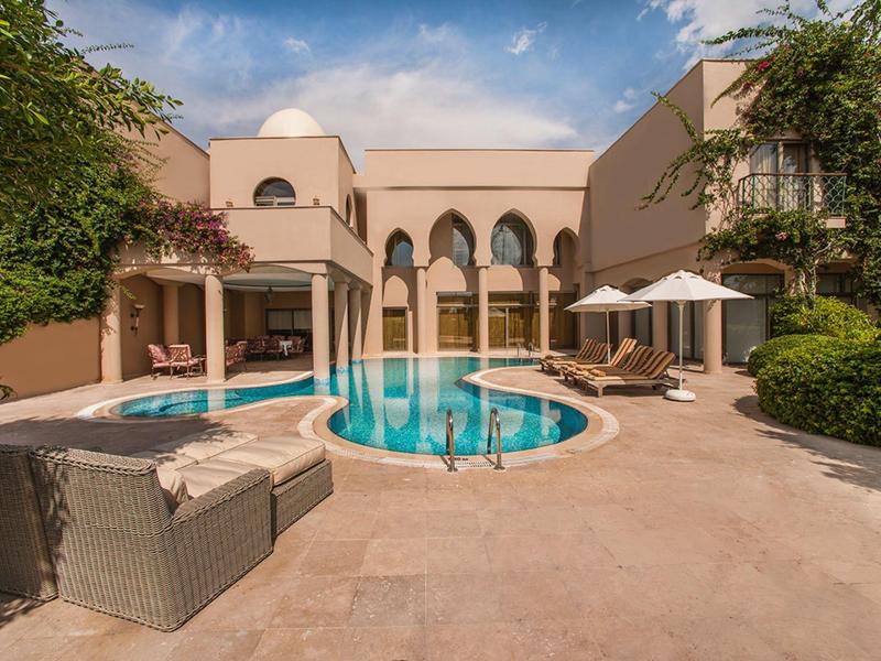 Basen hotelowy z okrągłymi i prostokątnymi basenami, miejscem do siedzenia i parasolami pod niebieskim niebem.