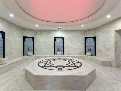 Großer, heller Hamam mit zentralem Steintisch, Marmwänden, Nischen und rosa Deckenbeleuchtung.