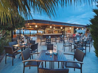 Außenbereich eines Restaurants mit Tischen, Stühlen, Beleuchtung und Palmen bei Abenddämmerung.