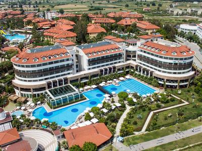 Großes Hotel mit roten Dächern, mehreren Pools, grünen Gärten und angrenzenden Wohngebäuden.