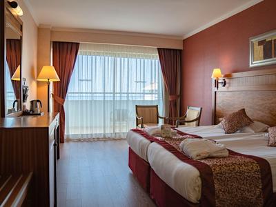 Camera d'hotel con letto matrimoniale, due poltrone, tende e balcone con vista mare.
