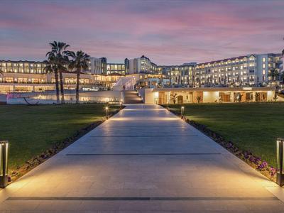 Lit pathway leads to modern hotel building at sunset with palm trees and green lawn.