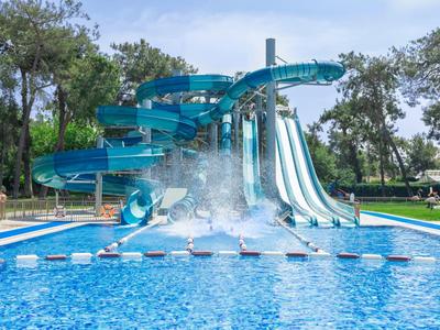 Meerdere glijbanen leiden naar een groot zwembad omringd door bomen en zonnige lucht.