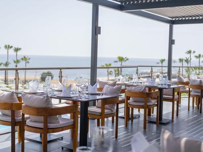 Otevřená restaurace s dřevěnými stoly a výhledem na moře za slunečného dne.