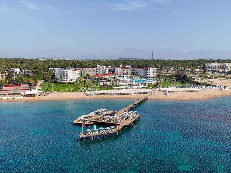 Wide wooden pier with restaurant over clear sea near sandy beach and hotel buildings.