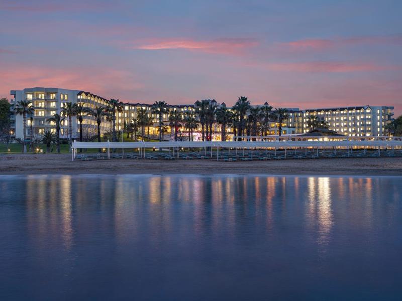Large hotel building with illuminated windows by calm water at sunset.