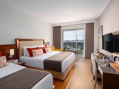 Světý hotelový pokoj se dvěma postelemi, TV, psacím stolem a balkonem s výhledem na městské budovy.