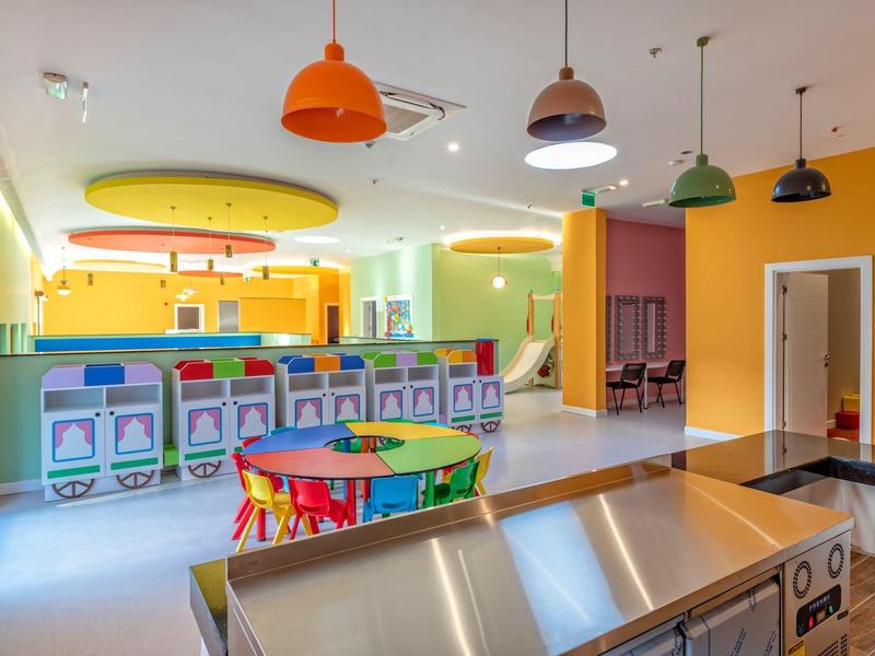 Jasná dětská herna s barevnými stoly, židlemi a arkádovými automaty pod závěsnými světly.