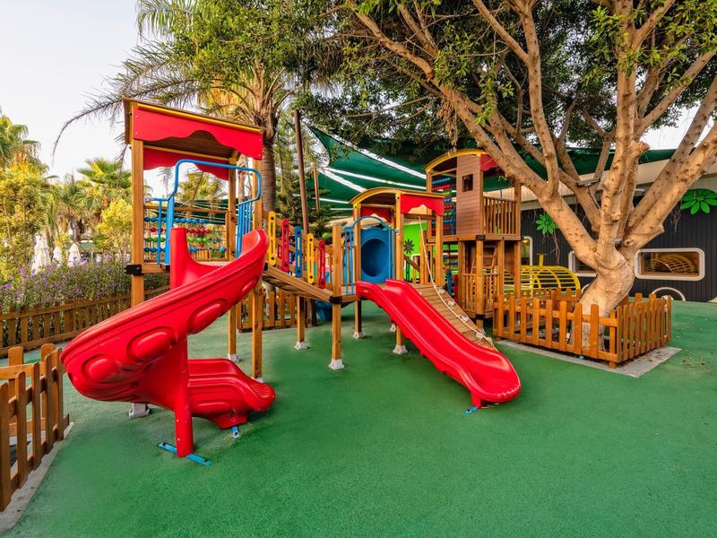 Barevné venkovní hřiště se skluzavkami a lezeckými konstrukcemi obklopené stromy a ploty.