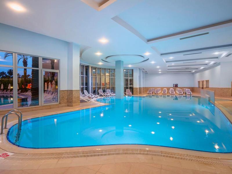 Vnitřní bazén s průzračnou modrou vodou, lehátky a velkými okny s výhledem na venkovní prostor.