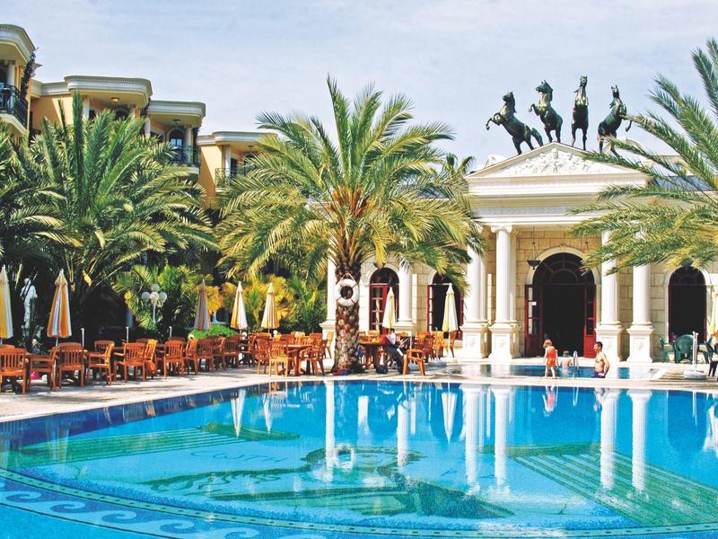Gran piscina de hotel rodeada de palmeras y estatuas, con mesas y sillas a los lados.