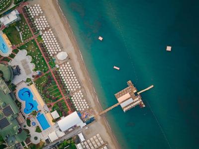 Vogelperspektive eines Strandes mit Liegen, Pools, grünen Pflanzen und einem langen Pier im blauen Meer.