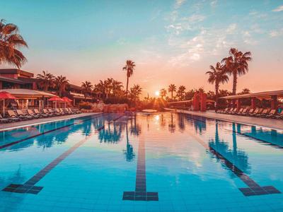 Ein großes Schwimmbecken mit blauen Fliesen, umgeben von Palmen und Liegestühlen bei Sonnenuntergang.