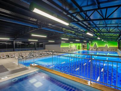 Hallenbad mit beleuchtetem Schwimmbecken, blauen Fliesen und Holzgeländer im Vordergrund.