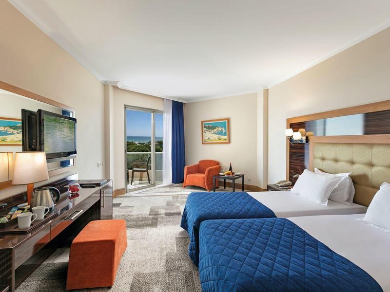 Helles Hotelzimmer mit zwei Betten, blauen Decken, orangefarbenen Möbeln und Balkon mit Meerblick.