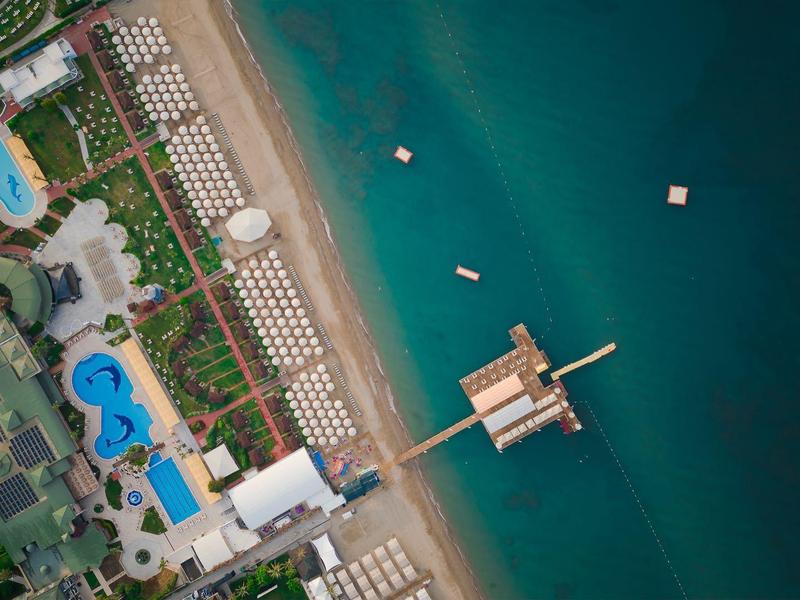 Vogelperspektive eines Strandes mit Liegen, Pools, grünen Pflanzen und einem langen Pier im blauen Meer.