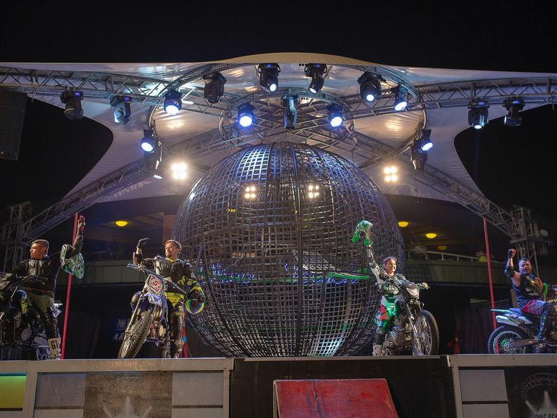 Bühne mit großem, beleuchtetem Globus und vier Personen auf Motorrädern bei Nacht.