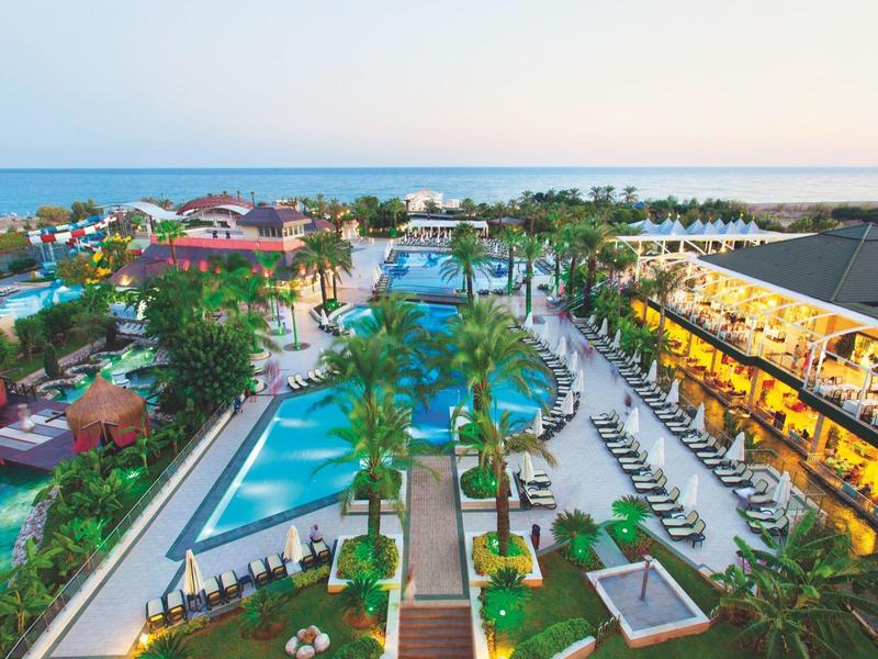 Moderner Hotelkomplex mit mehreren Pools, Liegestühlen und Blick auf das Meer bei Sonnenuntergang.