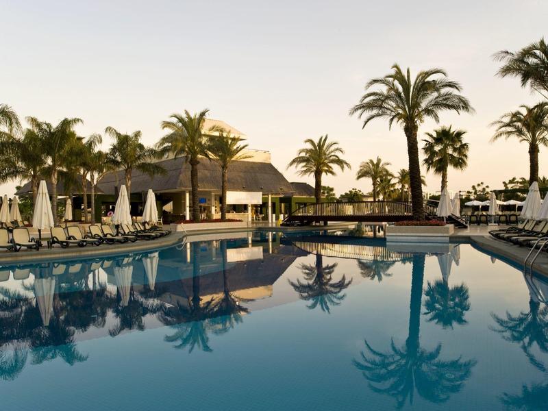 Großer Hotelpool mit Liegestühlen und Palmen bei Sonnenuntergang.