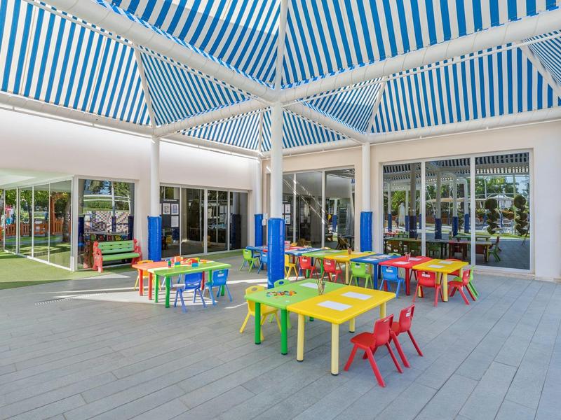 Bunter Kinderspielbereich mit Tischen und Stühlen unter einem gestreiften, überdachten Bereich.