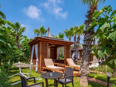 Wooden cabin with two lounge chairs in tropical garden with palm trees under clear sky.