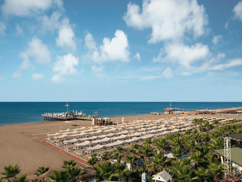 Vue d'une plage de sable avec des transats, des palmiers et une jetée sous un ciel bleu.