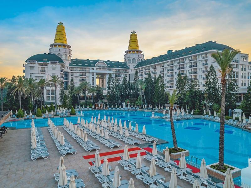 Grand piscine d'hôtel avec de nombreuses chaises longues et un bâtiment imposant en arrière-plan au coucher du soleil.
