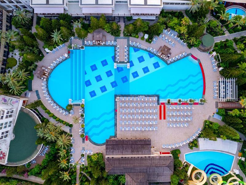 Vue aérienne d'une grande piscine bleue avec des transats et des arbres et bâtiments environnants.