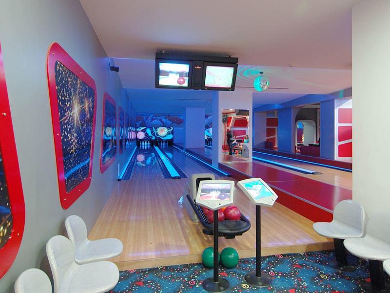 Allée de bowling moderne avec tapis coloré, chaises blanches et écrans lumineux.