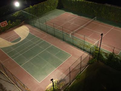 Zwei beleuchtete Tennisplätze mit grüner Umrandung und Netz, umgeben von Hecken in der Nacht.