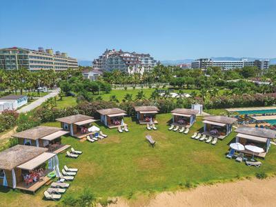 Grüne Rasenfläche mit Cabanas und Liegestühlen vor modernen Hotelgebäuden unter blauem Himmel.