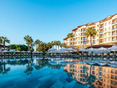 Moderner Hotelkomplex mit Palmen und Sonnenschirmen, daneben ein großer, klarer Pool.