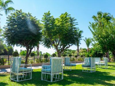 Grüne Wiese mit weißen Stühlen unter großen Bäumen und blauem Himmel im Gartenhotel.