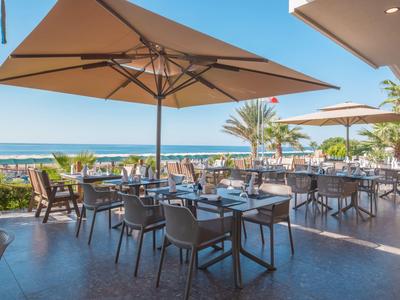 Terrassenrestaurant mit Tischen, Stühlen und Sonnenschirmen, mit Meerblick und Palmen.