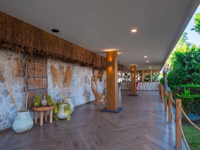 Überdachter Hotelflur mit Natursteinwand, Holzsäulen und grüner Umgebung neben Geländer.