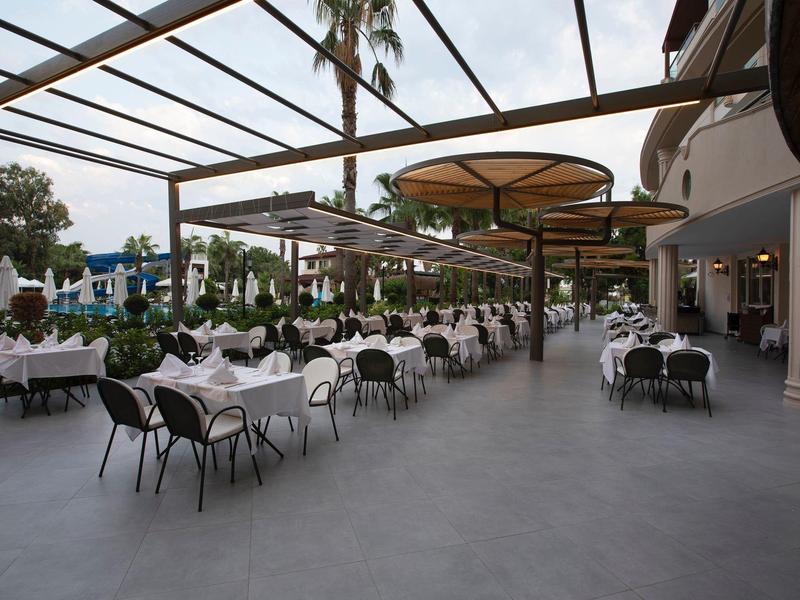 Amplia terraza con mesas cubiertas y sillas bajo toldos, rodeada de plantas y palmeras.