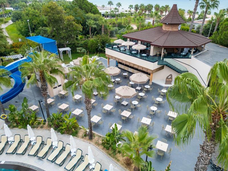 Amplia zona de piscina en hotel con tumbonas, mesas, palmeras y restaurante central con área cubierta.